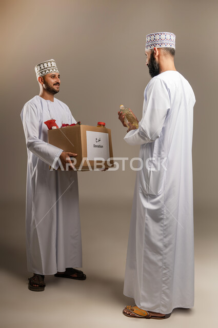 التقرب إلى الله بالعطاء للمحتاجين، المساعدة والتبرع لمشروع السلة الخيرية، بورتريه لرجل عربي خليجي عماني يرتدي الدشداشة والكمة يقوم بتقديم صندوق من المواد الغذائية لأحد المتعففين، مبادرة إنسانية في سلطنة عمان، توفير الطعام للفقراء، الجسم بالطول الكامل، خلفية بيج