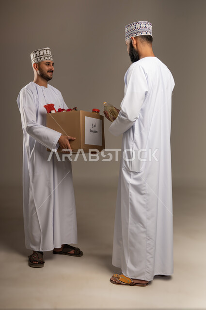 توفير الطعام للفقراء، التقرب إلى الله بالعطاء للمحتاجين، المساعدة والتبرع لمشروع السلة الخيرية، بورتريه لرجل عربي خليجي عماني يرتدي الدشداشة والكمة يقوم بتقديم صندوق من المواد الغذائية لأحد المتعففين، مبادرة إنسانية في سلطنة عمان، الجسم بالطول الكامل، خلفية بيج