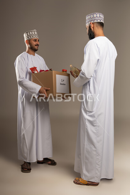 التقرب إلى الله بالعطاء للمحتاجين، المساعدة والتبرع لمشروع السلة الخيرية، بورتريه لرجل عربي خليجي عماني يرتدي الدشداشة والكمة يقوم بتقديم صندوق من المواد الغذائية لأحد المتعففين، مبادرة إنسانية في سلطنة عمان، توفير الطعام للفقراء، الجسم بالطول الكامل، خلفية بيج