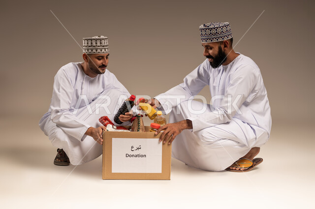 المساعدة والتبرع لمشروع السلة الخيرية، بورتريه لرجلين عربيين خليجيين عمانيين يرتديان الدشداشة والكمة يقومان بتجهيز صندوق من المواد الغذائية، مبادرة إنسانية في سلطنة عمان، توفير الطعام للفقراء، التقرب إلى الله بالعطاء للمحتاجين، خلفية بيج