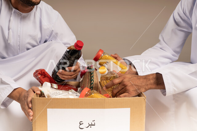 توفير الطعام للفقراء، المساعدة والتبرع لمشروع السلة الخيرية، بورتريه مقرب لرجلين عربيين خليجيين عمانيين يرتديان الدشداشة يقومان بتجهيز صندوق من المواد الغذائية، مبادرة إنسانية في سلطنة عمان، التقرب إلى الله بالعطاء للمحتاجين، خلفية بيج