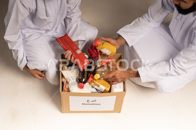 مبادرة إنسانية في سلطنة عمان، توفير الطعام للفقراء، المساعدة والتبرع لمشروع السلة الخيرية، بورتريه مقرب لرجلين عربيين خليجيين عمانيين يرتديان الدشداشة يقومان بتجهيز صندوق من المواد الغذائية، التقرب إلى الله بالعطاء للمحتاجين، خلفية بيج