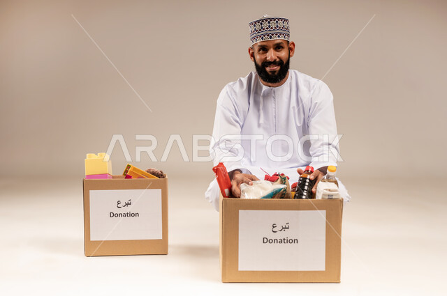 توفير الطعام للفقراء، مفهوم المساعدة والتبرع لمشروع السلة الخيرية، بورتريه لرجل عربي خليجي عماني يرتدي الدشداشة والكمة يقوم بتجهيز صندوق من المواد الغذائية، جمعية ومبادرة إنسانية في سلطنة عمان، التقرب إلى الله بالعطاء والصدقة للمحتاجين، خلفية بيج