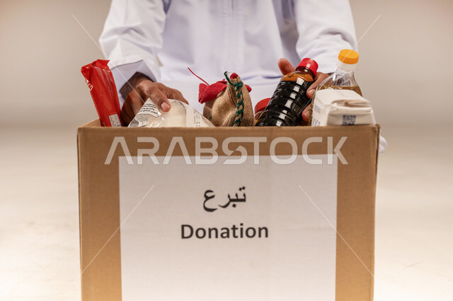 مفهوم المساعدة والتبرع لمشروع السلة الخيرية، بورتريه مقرب لرجل عربي خليجي عماني يرتدي الدشداشة يقوم بتجهيز صندوق من المواد الغذائية، جمعية ومبادرة إنسانية في سلطنة عمان، توفير الطعام للفقراء، التقرب إلى الله بالعطاء والصدقة للمحتاجين، خلفية بيج
