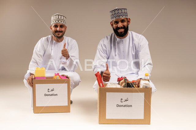 علامة الجودة والامتياز، توفير المستلزمات الضرورية للفقراء، مفهوم المساعدة والتبرع، بورتريه لرجلين عربيين خليجيين عمانيين يرتديان الدشداشة والكمة يقومان بتجهيز صناديق التبرعات يرفعان إصبع الإبهام بتعابير الاعجاب، تقديم الألعاب للأطفال المحتاجين، خلفية بيج