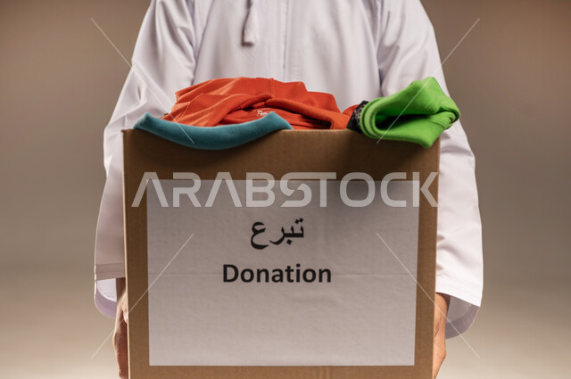 توفير المستلزمات الضرورية للفقراء، جمعية خيرية ومبادرة انسانية في سلطنة عمان، تأمين الملابس وإهدائها للمحتاجين، بورتريه مقرب لرجل عربي خليجي عماني يرتدي الدشداشة يحمل صندوق من الثياب، مفهوم المساعدة والتبرع، خلفية بيج