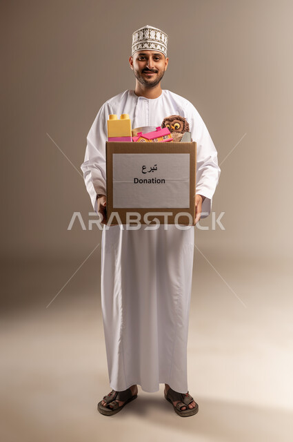 توفير وسائل التسلية والترفيه، بورتريه لرجل عربي خليجي عماني يرتدي الدشداشة والكمة يحمل صندوق مليء بألعاب الأطفال ينظر إلى الكاميرا بإيماءات السرور والسعادة، جمعية خيرية ومبادرة انسانية في سلطنة عمان، مفهوم المساعدة والتبرع، الجسم بالطول الكامل، خلفية بيج