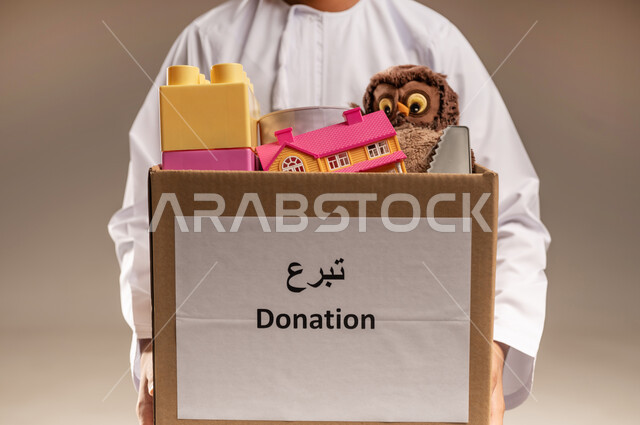 جمعية خيرية ومبادرة انسانية في سلطنة عمان، توفير وسائل التسلية والترفيه، بورتريه مقرب لرجل عربي خليجي عماني يرتدي الدشداشة يحمل صندوق مليء بألعاب الأطفال، مفهوم المساعدة والتبرع، خلفية بيج