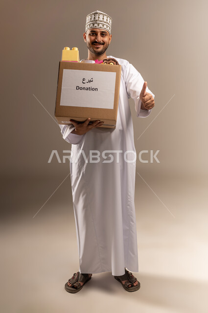 علامة الجودة والامتياز، توفير وسائل التسلية والترفيه، بورتريه لرجل عربي خليجي عماني يرتدي الدشداشة والكمة يحمل صندوق مليء بألعاب الأطفال يرفع إصبع الإبهام ينظر إلى الكاميرا بإيماءات الإعجاب، مفهوم المساعدة والتبرع، الجسم بالطول الكامل، خلفية بيج