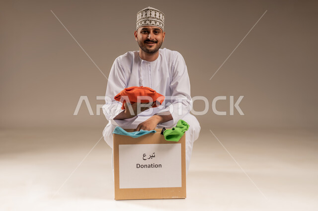 مفهوم المساعدة والتبرع، بورتريه لرجل عربي خليجي عماني يرتدي الدشداشة والكمة يقوم بتجهيز صندوق من الثياب ينظر إلى الكاميرا بإيماءات السرور، تأمين الملابس وإهدائها للمحتاجين، توفير المستلزمات الضرورية للفقراء، جمعية خيرية ومبادرة انسانية في سلطنة عمان، خلفية بيج