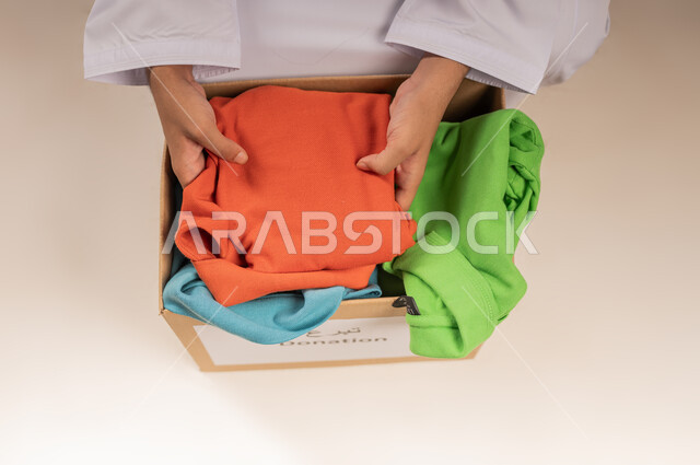 توفير المستلزمات الضرورية للفقراء، جمعية خيرية ومبادرة انسانية في سلطنة عمان، تأمين الملابس وإهدائها للمحتاجين، بورتريه مقرب لرجل عربي خليجي عماني يرتدي الدشداشة يقوم بتجهيز صندوق من الثياب، مفهوم المساعدة والتبرع، خلفية بيج