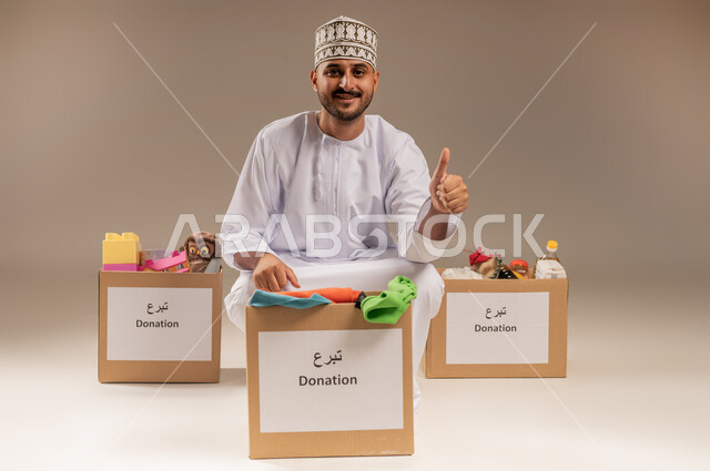 علامة الجودة والامتياز، توفير المستلزمات الضرورية للفقراء، مفهوم المساعدة والتبرع، بورتريه لرجل عربي خليجي عماني يرتدي الدشداشة والكمة يقوم بتجهيز صناديق التبرعات، رفع إصبع الإبهام بإيماءات الإعجاب، تقديم الألعاب والملابس للأطفال المحتاجين، خلفية بيج