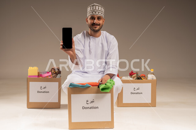 عرض شاشة سوداء فارغة، توفير المستلزمات الضرورية للفقراء، مفهوم المساعدة والتبرع، بورتريه لرجل عربي خليجي عماني يرتدي الدشداشة والكمة يمسك الهاتف المحمول ينظر إلى الكاميرا بإيماءات السرور، استخدام جهاز تقني حديث، تقديم الألعاب والملابس للأطفال المحتاجين، خلفية بيج
