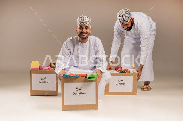 مبادرة إنسانية في سلطنة عمان، توفير المستلزمات الضرورية للفقراء، مفهوم المساعدة والتبرع، بورتريه لرجلين عربيين خليجيين عمانيين يرتديان الدشداشة والكمة يقومان بتجهيز صناديق التبرعات، تقديم الألعاب والملابس للأطفال المحتاجين، خلفية بيج