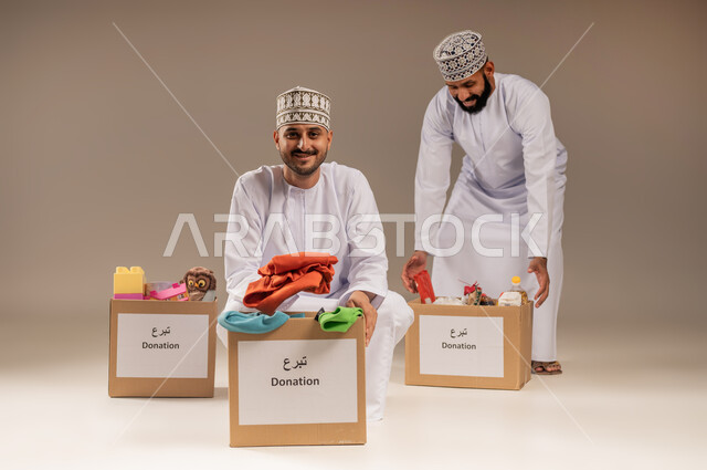 توفير المستلزمات الضرورية للفقراء، مفهوم المساعدة والتبرع، بورتريه لرجلين عربيين خليجيين عمانيين يرتديان الدشداشة والكمة يقومان بتجهيز صناديق التبرعات، مبادرة إنسانية في سلطنة عمان، تقديم الألعاب والملابس للأطفال المحتاجين، خلفية بيج