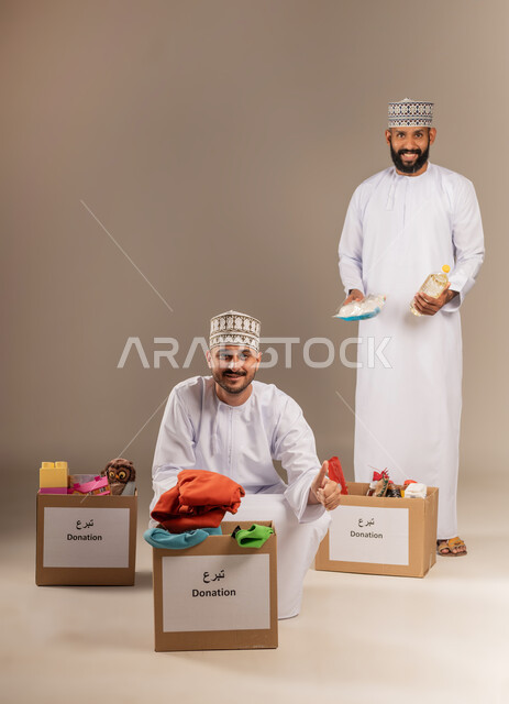 تقديم الألعاب والملابس للأطفال المحتاجين، توفير المستلزمات الضرورية للفقراء، مفهوم المساعدة والتبرع، بورتريه لرجلين عربيين خليجيين عمانيين يرتديان الدشداشة والكمة يقومان بتجهيز صناديق التبرعات، مبادرة إنسانية في سلطنة عمان، خلفية بيج