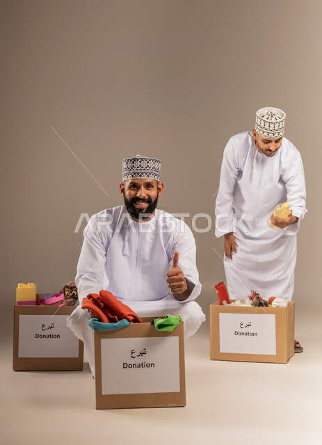 علامة الجودة والامتياز، توفير المستلزمات الضرورية للفقراء، مفهوم المساعدة والتبرع، بورتريه لرجلين عربيين خليجيين عمانيين يرتديان الدشداشة والكمة يقومان بتجهيز صناديق التبرعات، رفع إصبع الإبهام بتعابير الاعجاب، تقديم الألعاب والملابس للأطفال المحتاجين، خلفية بيج