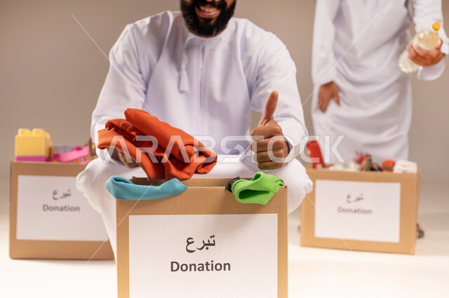 رفع إصبع الإبهام بإيماءات الإعجاب، علامة الجودة والامتياز، توفير المستلزمات الضرورية للفقراء، مفهوم المساعدة والتبرع، بورتريه مقرب لرجلين عربيين خليجيين عمانيين يرتديان الدشداشة يقومان بتجهيز صناديق التبرعات، تقديم الألعاب والملابس للأطفال المحتاجين، خلفية بيج