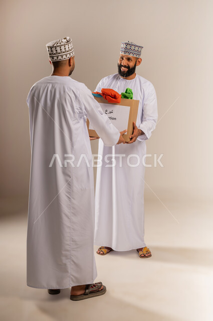توفير المستلزمات الضرورية للفقراء، مفهوم المساعدة والتبرع، بورتريه لرجل عربي خليجي عماني يرتدي الدشداشة والكمة يقوم بتقديم طرد من الثياب لشخص آخر، جمعية خيرية ومبادرة انسانية في سلطنة عمان، تأمين الملابس وإهدائها للمحتاجين، الجسم بالطول الكامل، خلفية بيج