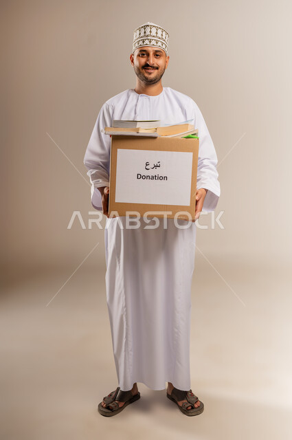 مفهوم المساعدة والتبرع، توفير المؤلفات التعليمية للمحتاجين، بورتريه لرجل عربي خليجي عماني يرتدي الدشداشة والكمة يحمل صندوق مليء بالكتب ينظر إلى الكاميرا بإيماءات السرور والسعادة، جمعية خيرية ومبادرة انسانية في سلطنة عمان، الجسم بالطول الكامل، خلفية بيج