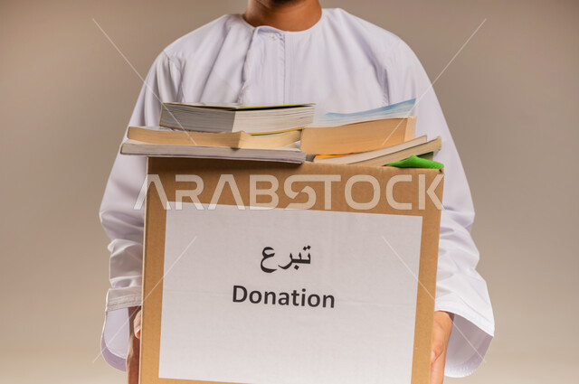 جمعية خيرية ومبادرة انسانية في سلطنة عمان، مفهوم المساعدة والتبرع، توفير المؤلفات التعليمية للمحتاجين، بورتريه مقرب لرجل عربي خليجي عماني يرتدي الدشداشة يحمل صندوق مليء بالكتب، خلفية بيج