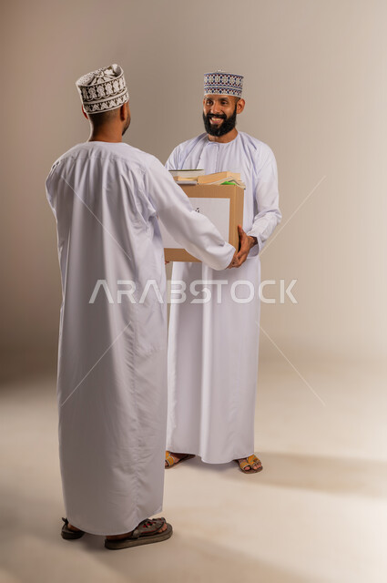 توفير المؤلفات التعليمية المفيدة للمحتاجين، بورتريه لرجل عربي خليجي عماني يرتدي الدشداشة والكمة يحمل صندوق مليء بالكتب الفائضة يقوم بتقديمه إلى شاب آخر، جمعية خيرية ومبادرة انسانية في سلطنة عمان، مفهوم المساعدة والتبرع، الجسم بالطول الكامل، خلفية بيج