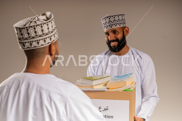مفهوم المساعدة والتبرع، توفير المؤلفات التعليمية المفيدة للمحتاجين، بورتريه مقرب لرجل عربي خليجي عماني يرتدي الدشداشة والكمة يحمل صندوق مليء بالكتب الفائضة يقوم بتقديمه إلى شاب آخر، جمعية خيرية ومبادرة انسانية في سلطنة عمان، خلفية بيج