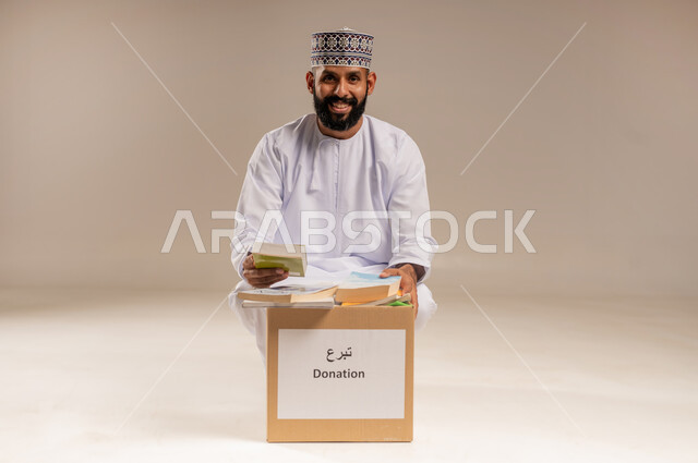 جمعية خيرية ومبادرة انسانية في سلطنة عمان، مفهوم المساعدة والتبرع، توفير المؤلفات التعليمية للمحتاجين، بورتريه لرجل عربي خليجي عماني يرتدي الدشداشة والكمة يقوم بتجهيز صندوق تبرعات من الكتب الفائضة ينظر إلى الكاميرا بإيماءات السرور، خلفية بيج
