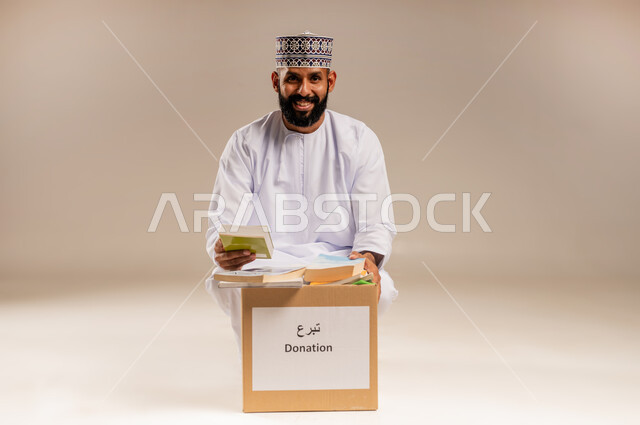 مفهوم المساعدة والتبرع، توفير المؤلفات التعليمية للمحتاجين، بورتريه لرجل عربي خليجي عماني يرتدي الدشداشة والكمة يقوم بتجهيز صندوق تبرعات من الكتب الفائضة ينظر إلى الكاميرا بإيماءات السرور، جمعية خيرية ومبادرة انسانية في سلطنة عمان، خلفية بيج
