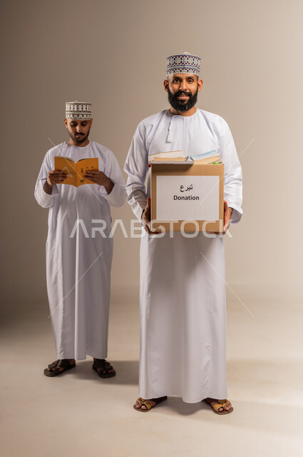 توفير المؤلفات التعليمية المفيدة للمحتاجين، بورتريه لرجل عربي خليجي عماني يرتدي الدشداشة والكمة يحمل صندوق مليء بالكتب الفائضة يقف برفقة شاب يتصفح كتاب، جمعية خيرية ومبادرة انسانية في سلطنة عمان، مفهوم المساعدة والتبرع، الجسم بالطول الكامل، خلفية بيج