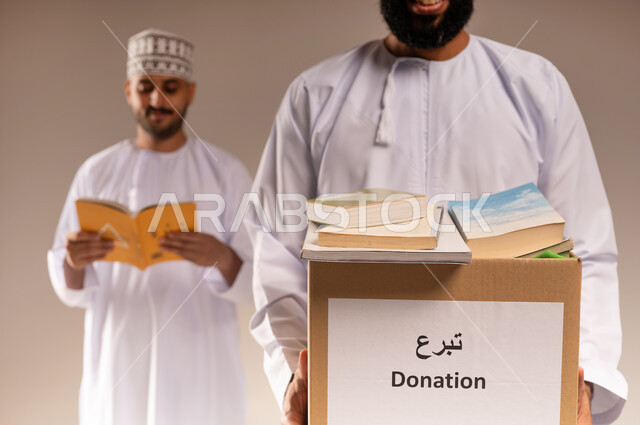 مفهوم المساعدة والتبرع، توفير المؤلفات التعليمية المفيدة للمحتاجين، بورتريه لرجل عربي خليجي عماني يرتدي الدشداشة يحمل صندوق مليء بالكتب الفائضة يقف برفقة شاب يتصفح كتاب، جمعية خيرية ومبادرة انسانية في سلطنة عمان، خلفية بيج
