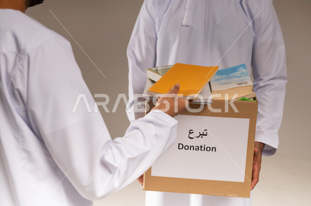 توفير المؤلفات التعليمية المفيدة للمحتاجين، بورتريه لرجل عربي خليجي عماني يرتدي الدشداشة يحمل صندوق مليء بالكتب الفائضة يقوم بتقديمه إلى شاب آخر، جمعية خيرية ومبادرة انسانية في سلطنة عمان، مفهوم المساعدة والتبرع، خلفية بيج