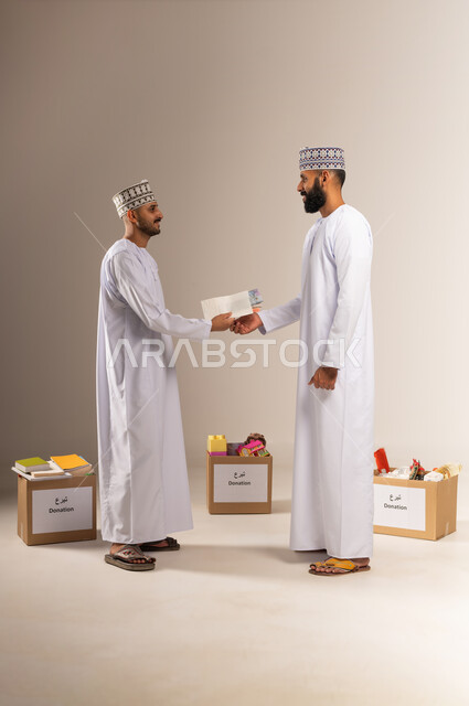 Giving zakat money to the needy, the concept of helping and donating, a portrait of an Arab Gulf Omani man wearing a dishdasha and a kummah holding an envelope of paper money that he is giving to another person, getting closer to God through giving and charity, a group of donation boxes on the ground, full-length body, beige background