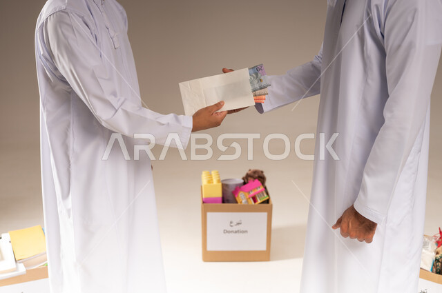 مفهوم المساعدة والتبرع، بورتريه مقرب لرجل عربي خليجي عماني يرتدي الدشداشة يحمل بيده ظرف من النقود الورقية يقوم بتقديمه لشخص آخر، التقرب إلى الله بالعطاء والصدقة، جمعية خيرية ومبادرة إنسانية في سلطنة عمان، إخراج زكاة المال للمحتاجين، خلفية بيج