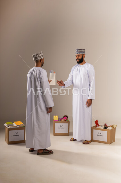 A group of donation boxes on the ground, the concept of helping and donating, a portrait of an Arab Gulf Omani man wearing a dishdasha and a kummah holding an envelope of paper money in his hand and giving it to another person, getting closer to God through giving and charity, giving zakat money to the needy, full body, beige background