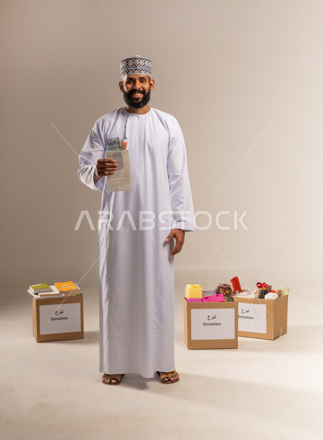 إخراج زكاة المال للمحتاجين، مفهوم المساعدة والتبرع، بورتريه لرجل عربي خليجي عماني يرتدي الدشداشة والكمة يقف أمام صناديق التبرعات يحمل بيده النقود الورقية ينظر إلى الكاميرا بإيماءات السرور، العبادة والتقرب إلى الله بالعطاء والصدقة، الجسم بالطول الكامل، خلفية بيج