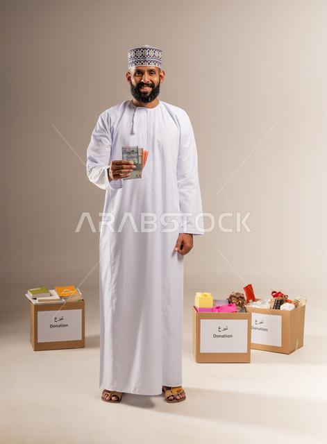 The concept of helping and donating, portrait of an Arab Gulf Omani man wearing a dishdasha and a kummah standing in front of donation boxes holding paper money in his hand looking at the camera with gestures of pleasure, worship and getting closer to God through giving and charity, giving zakat money to the needy, full body, beige background
