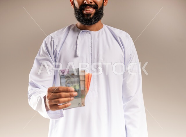 العبادة والتقرب إلى الله بالعطاء والصدقة، جمعية خيرية ومبادرة إنسانية في سلطنة عمان، مفهوم المساعدة والتبرع، بورتريه مقرب لرجل عربي خليجي عماني مبتسم يرتدي الدشداشة يحمل بيده النقود الورقية، إخراج زكاة المال للمحتاجين، خلفية بيج