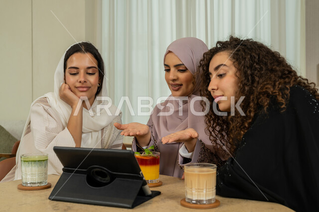 Browsing the internet on a tablet, a fun and acquaintance session, spending quality time with friends, developing social relationships, Arab Gulf Emirati female friends sitting at a table in a café, Saudi women doing a live broadcast on social media, a high-end coffee shop in the Emirates