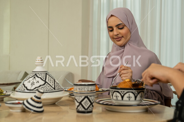 جلسة وناسة في عطلة نهاية الاسبوع, ايماءات السعادة والسرور، اجتماع الرفاق في مطعم فاخر بالامارات، قضاء وقت ممتع برفقة الأصدقاء، امرأة عربية خليجية اماراتية محجبة مبتسمة تجلس على طاولة طعام مليئة بالأطعمة والمأكولات الشهية مع صديقاتها، سيدات سعوديات يتناولن العشاء