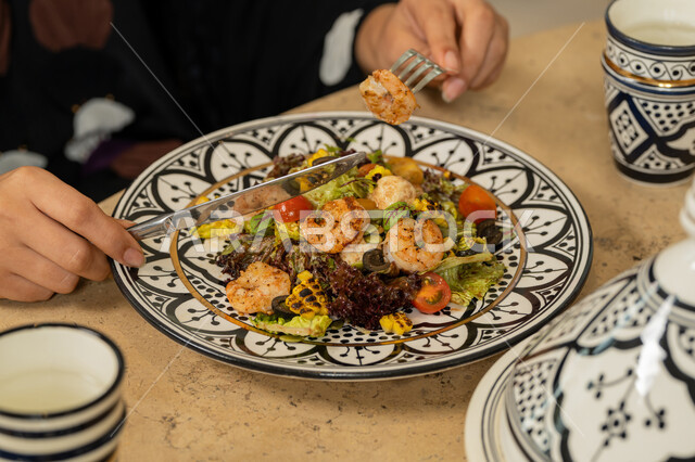 Delicious food and drinks, luxury restaurant in UAE, close-up of hands of an Emirati Gulf Arab woman holding a fork and knife and eating a plate of delicious shrimp, Saudi woman sitting at the dining table, having a good time on the weekend