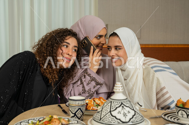 Making a phone call on a mobile phone, gestures of happiness and pleasure, friends meeting at a dining table in a fancy restaurant in the Emirates, spending a good time with friends, Arab Gulf Emirati female friends having dinner together outside, Saudi women listening to the conversation