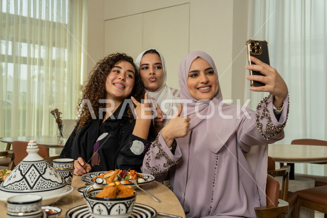 قضاء وقت ممتع برفقة الأصدقاء، التقاط صور السيلفي التذكارية، تجمع الرفاق على طاولة طعام في مطعم فاخر بالامارات، صديقات عربيات خليجيات اماراتيات يقومون بتناول العشاء معا في الخارج، سيدات سعوديات يستخدمون الهاتف المحمول، ايماءات السعادة والسرور
