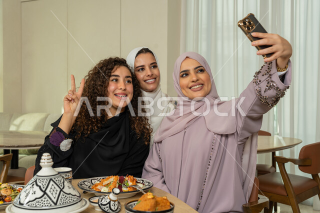 التقاط صور السيلفي التذكارية، قضاء وقت ممتع برفقة الأصدقاء، تجمع الرفاق على طاولة طعام في مطعم فاخر بالامارات، صديقات عربيات خليجيات اماراتيات يقومون بتناول العشاء معا في الخارج، سيدات سعوديات يستخدمون الهاتف المحمول، ايماءات السعادة والسرور