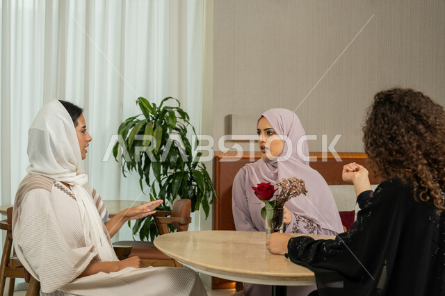Developing social relations, a fun and acquaintance session on a holiday, spending a good time chatting and socializing with friends, expressions of happiness and pleasure when gathering friends, a high-end coffee shop in the Emirates, Arab Gulf Emirati female friends sitting at a table in the café, Saudi women exchanging talk