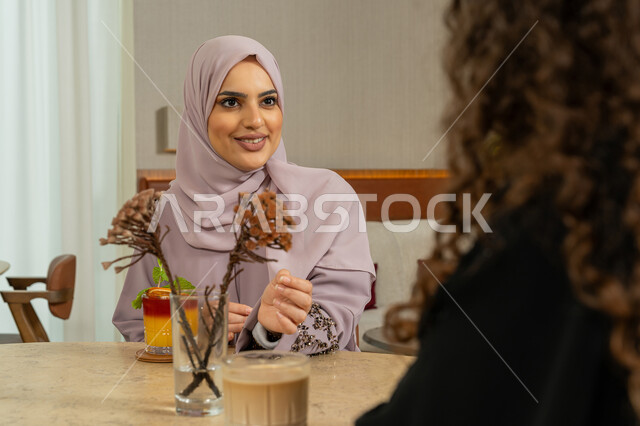 Expressions of happiness and pleasure with a gathering of friends, enjoying cold drinks, a pleasant session on a holiday, a high-end coffee shop in the Emirates, developing social relations, chatting with friends, a veiled Emirati Gulf Arab woman sitting with her friend in a café, two Saudi friends exchanging talk