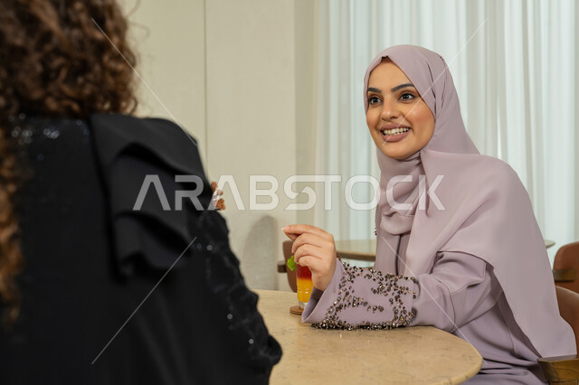 الاستمتاع بالمشروبات الباردة، جلسة وناسة يوم الاجازة، كوفي شوب راقي بالامارات، تطوير العلاقات الإجتماعية، التسامر والدردشة مع الأصدقاء، تعابير السعادة والسرور بلمة الرفاق، امرأة عربية خليجية اماراتية محجبة تجلس مع صديقتها في المقهى، صديقتان سعوديتان تتبادلان الحديث