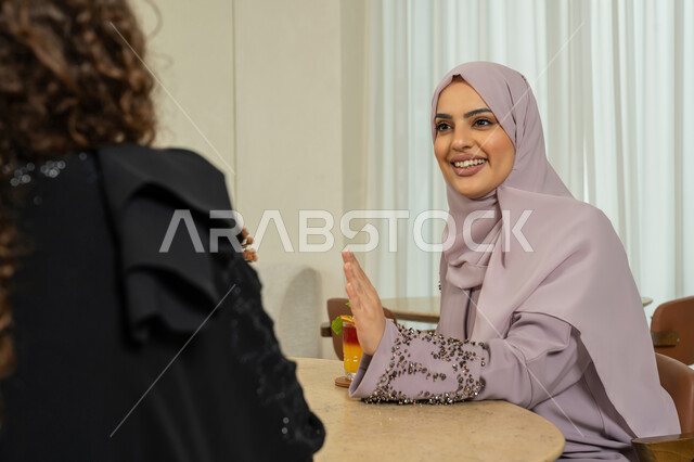 A fun session on a holiday, a classy coffee shop in the Emirates, developing social relations, chatting with friends, expressions of happiness and pleasure when gathering friends, enjoying cold drinks, a veiled Emirati Gulf Arab woman sitting with her friend in a café, two Saudi friends exchanging talk