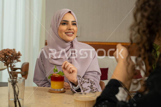تطوير العلاقات الإجتماعية، التسامر والدردشة مع الأصدقاء، تعابير السعادة والسرور بلمة الرفاق، الاستمتاع بالمشروبات الباردة، جلسة وناسة يوم الاجازة، كوفي شوب راقي بالامارات، امرأة عربية خليجية اماراتية محجبة تجلس مع صديقتها في المقهى، صديقتان سعوديتان تتبادلان الحديث