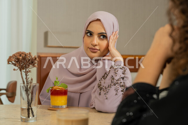 تعابير الاندماج والتركيز، جلسة وناسة وتعارف في يوم العطلة، التسامر والدردشة وقضاء وقت مع الأصدقاء، كوفي شوب راقي بالامارات، تطوير العلاقات الإجتماعية، امرأة عربية خليجية اماراتية محجبة تجلس مع رفاقها على طاولة في المقهى، سيدة سعودية تقوم بالاستماع والاصغاء